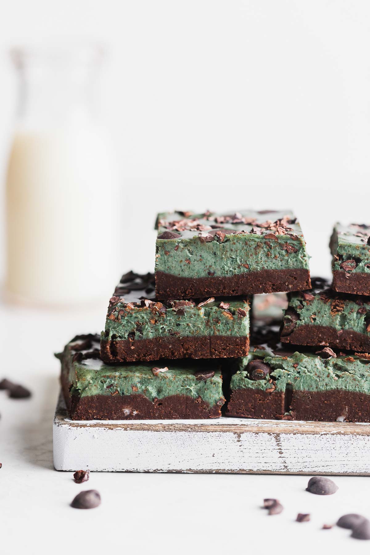 Paleo mint bars stacked up with milk in the back on a wooden cutting board.