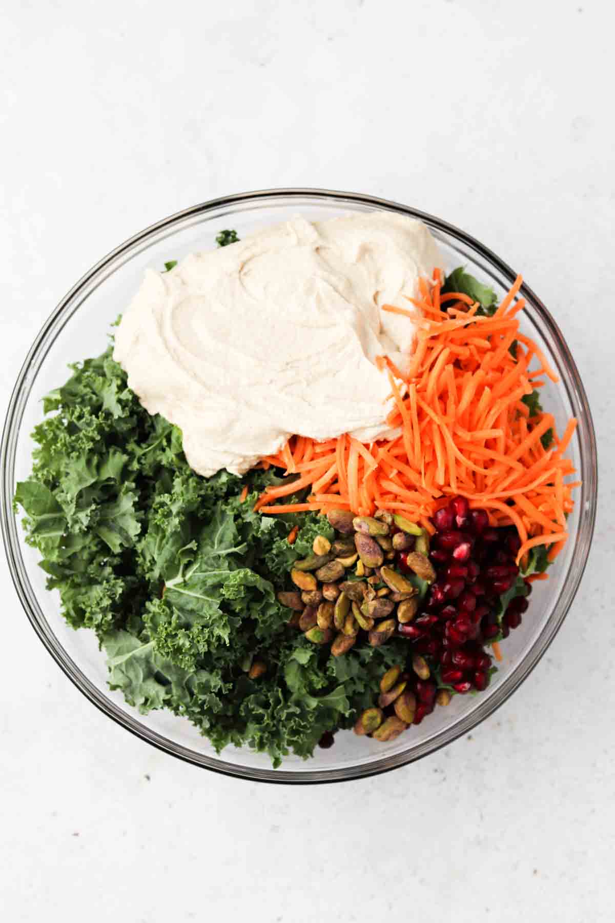 individual salad ingredients covering kale, carrots, tahini, nuts and more in a glass bowl.