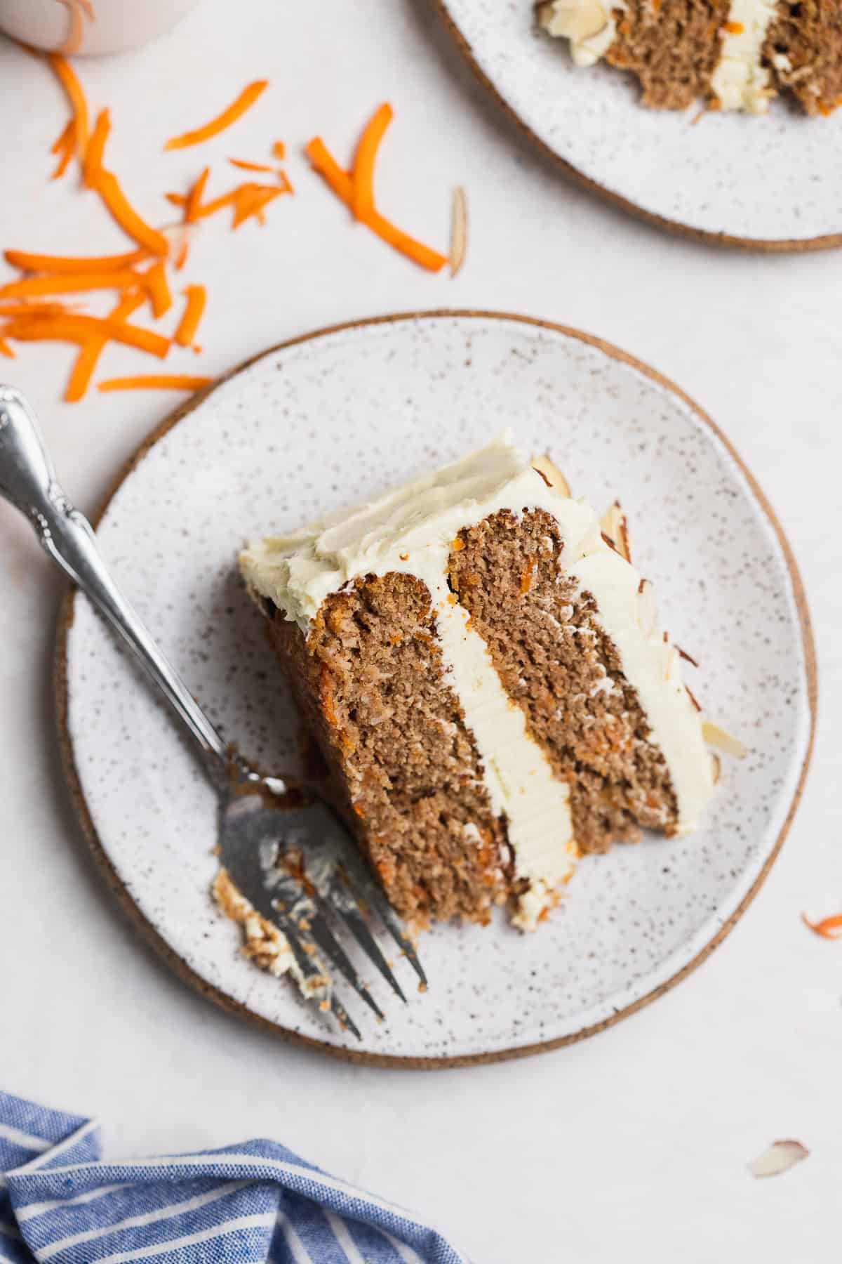 One slice of gluten free carrot cake on a plate with a fork with shredded carrot on the side. 
