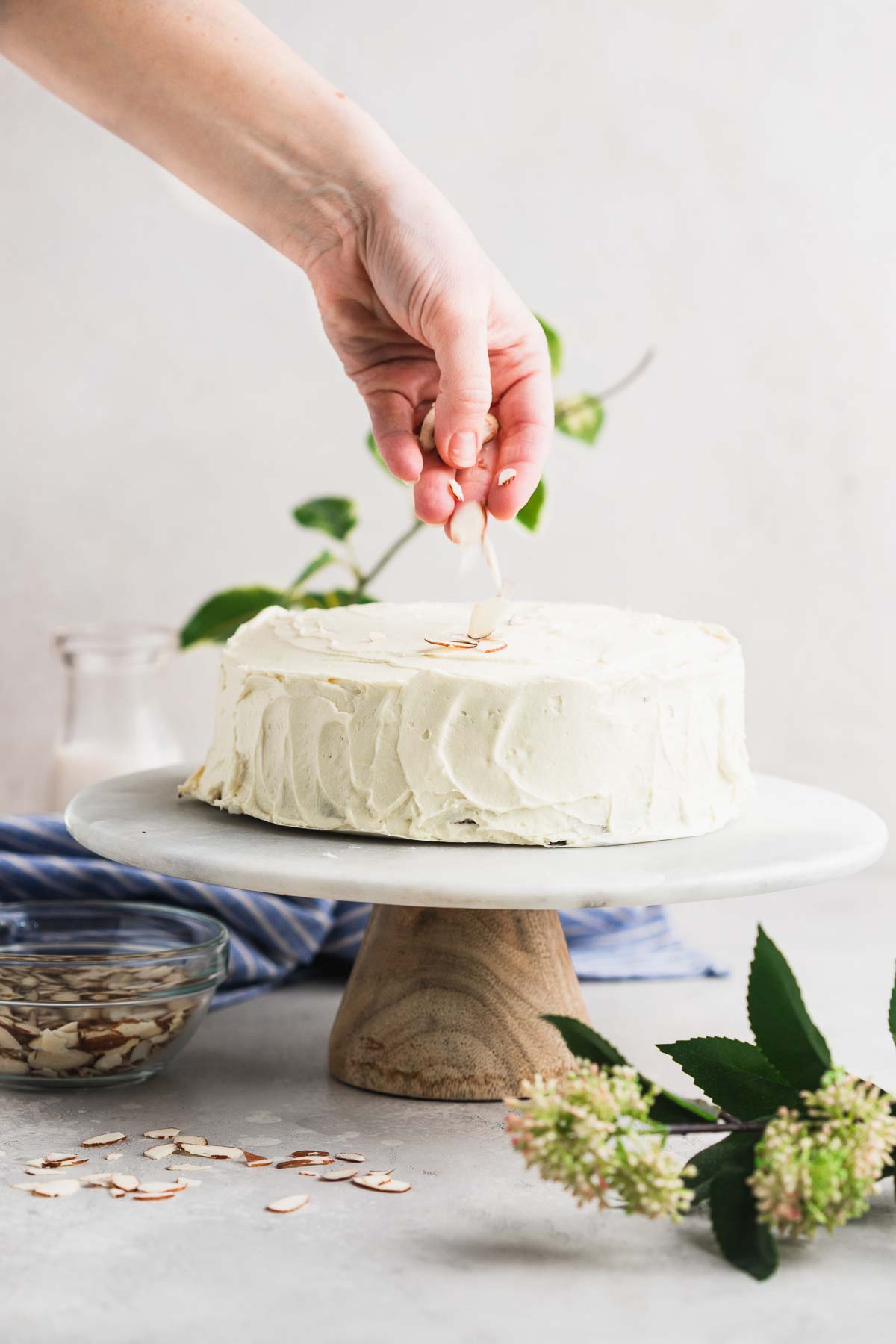 A hand sprinkling almond slivers on top of the cake on a cake stand.