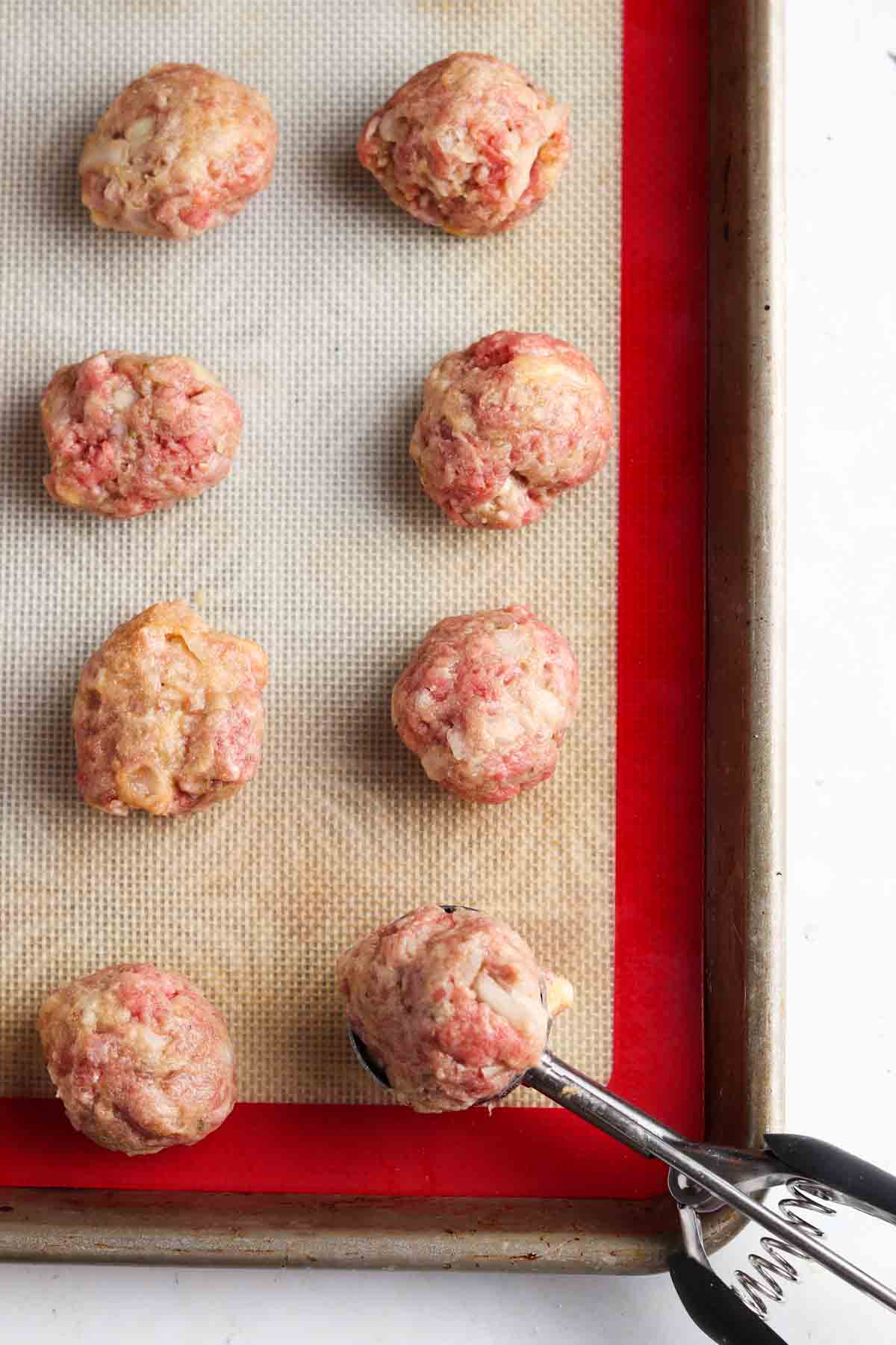 Raw ground beef meatballs on a baking tray with a cookie scooper.