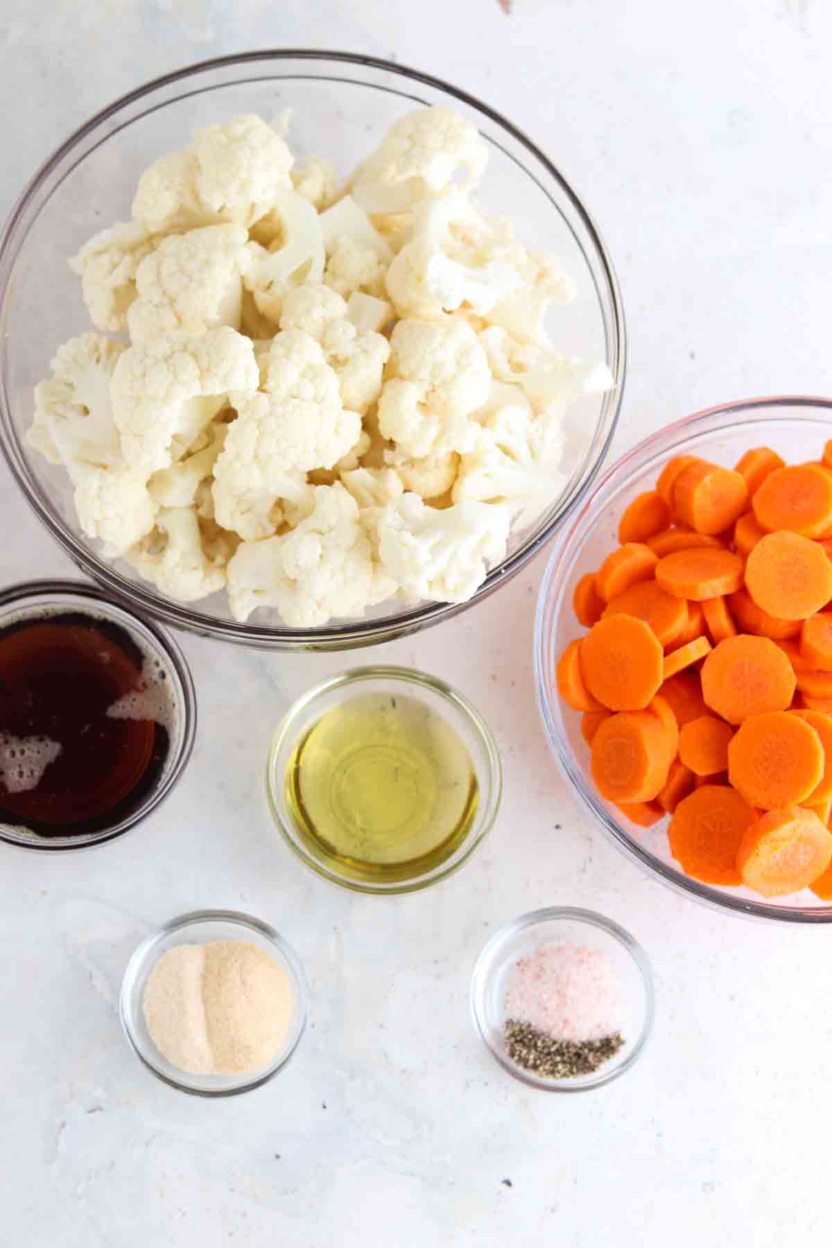 Carrots, cauliflower, seasoning, oil and maple syrup in small glass bowls on the counter. 