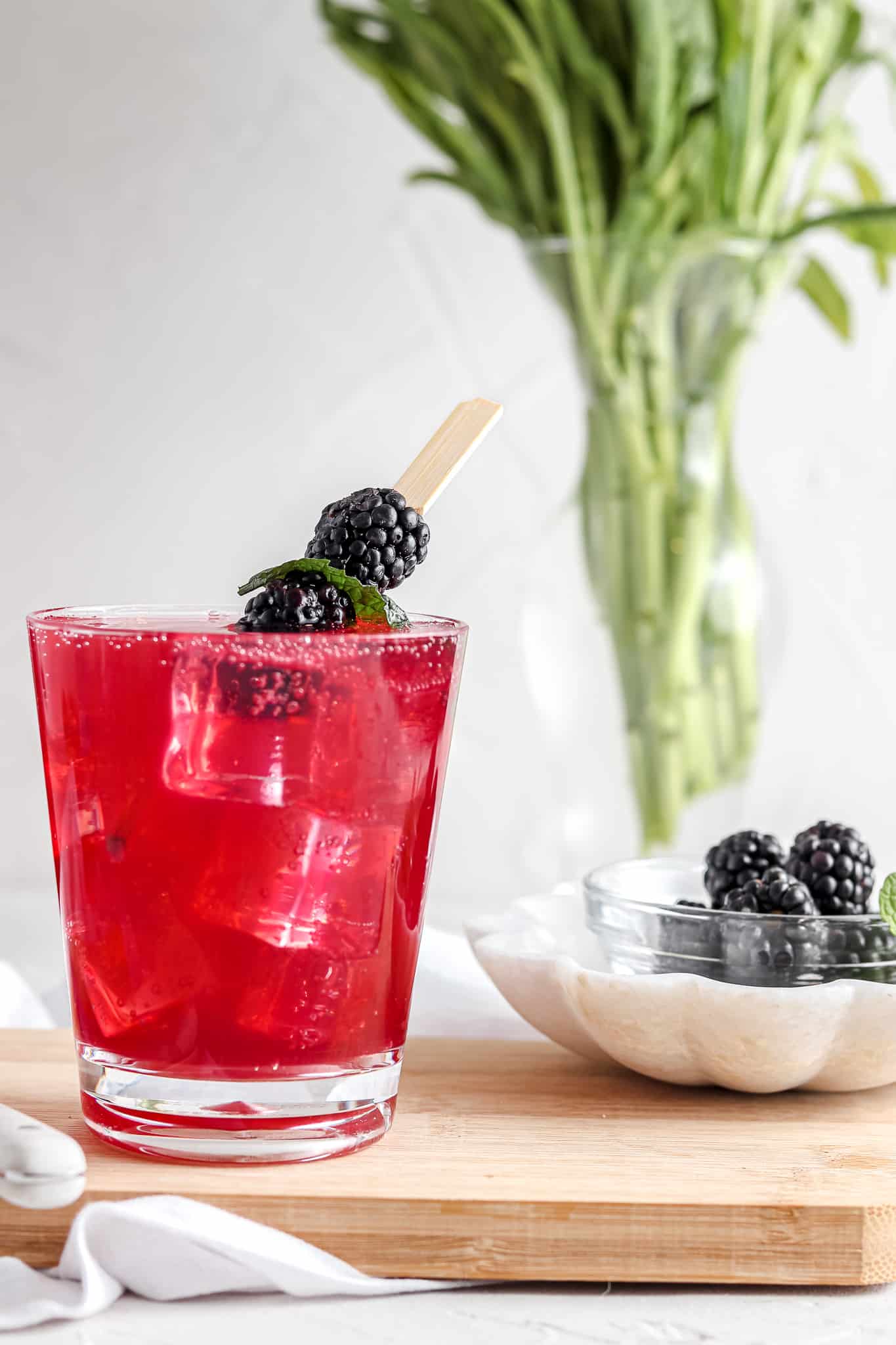 Blackberry mojito in a clear cup with fresh blackberries and mint on top sitting next to flowers in a vase. 