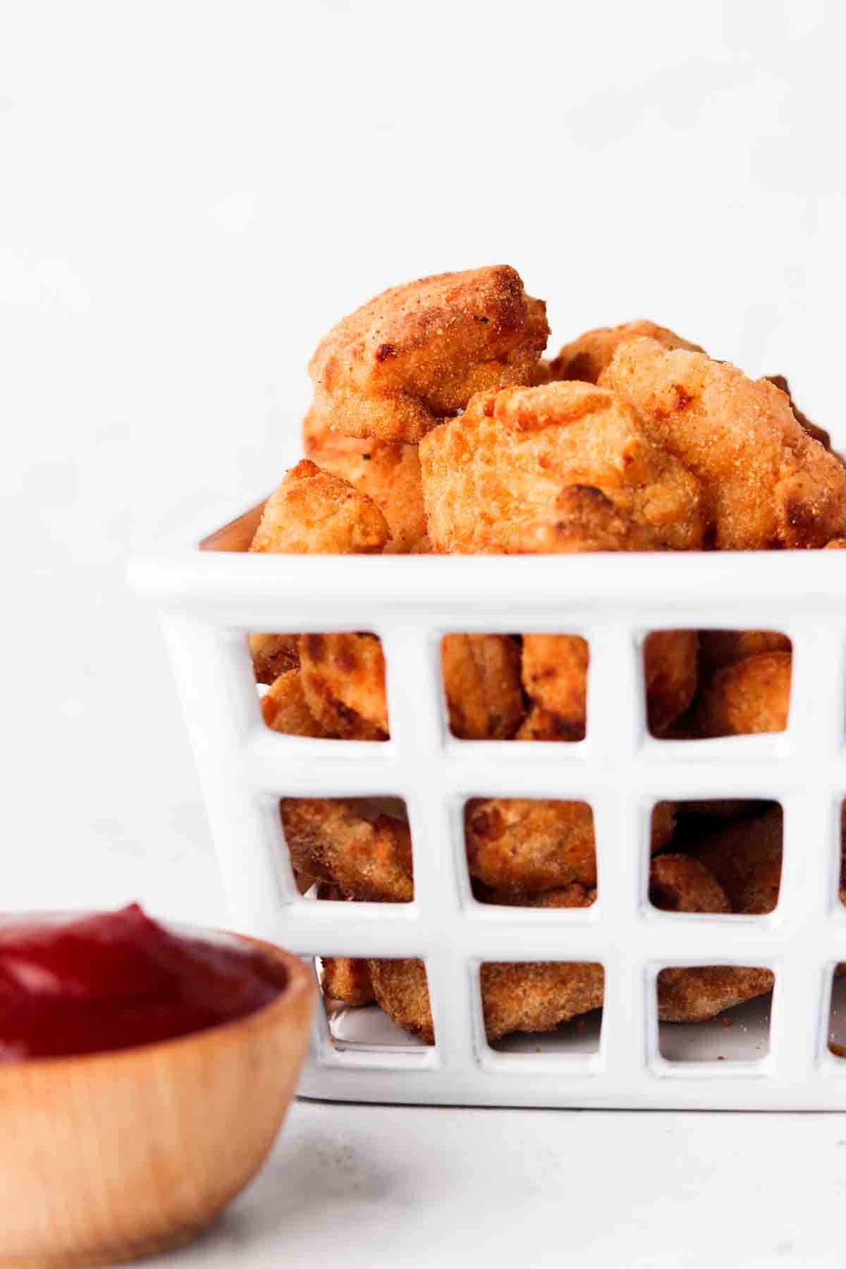 Crispy chicken nuggets in a white basket with a wooden bowl of ketchup next to it.