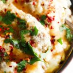 Close up photo of mashed potatoes in a bowl with gravy, parsley and red pepper flakes on top.