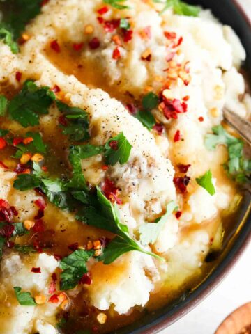Close up photo of mashed potatoes in a bowl with gravy, parsley and red pepper flakes on top.