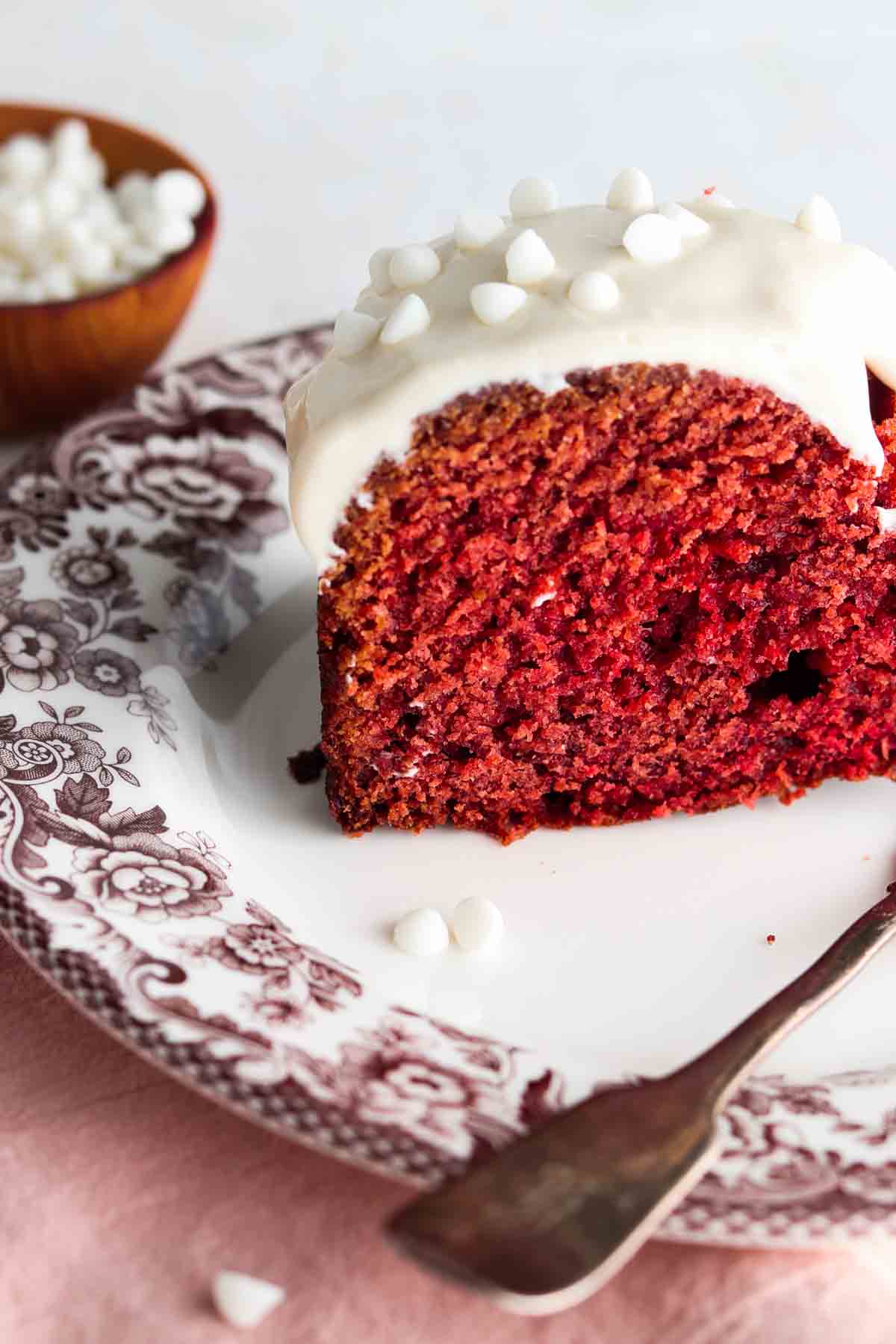 Slice of red velvet cake on a plate with white chocolate on top of the icing.
