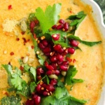 Pumpkin carrot soup in a scalloped bowl with parsley and pomegranate on the side.