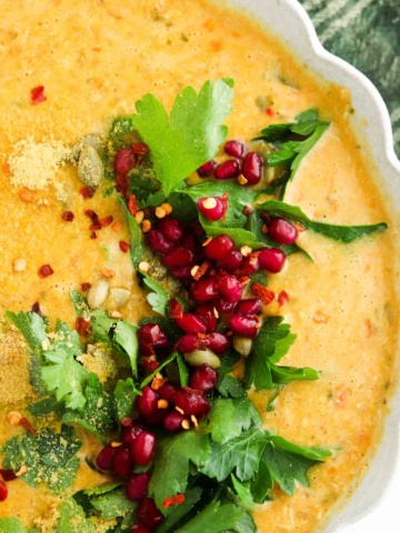 Pumpkin carrot soup in a scalloped bowl with parsley and pomegranate on the side.
