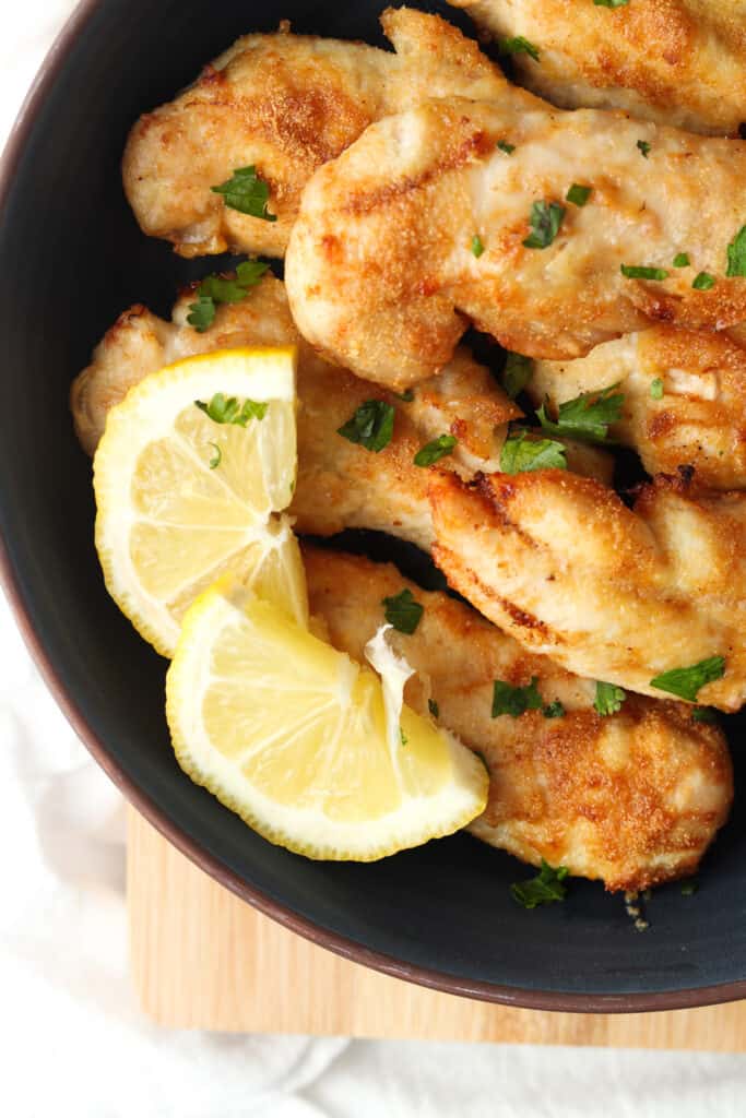 chicken tenders on a black plate with lemon slices and sprinkled with green herbs