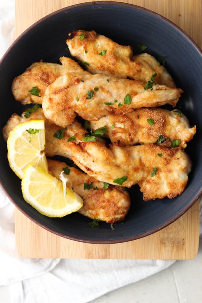 crispy chicken tenders in a black bowl with lemon wedges with green herb garnish 