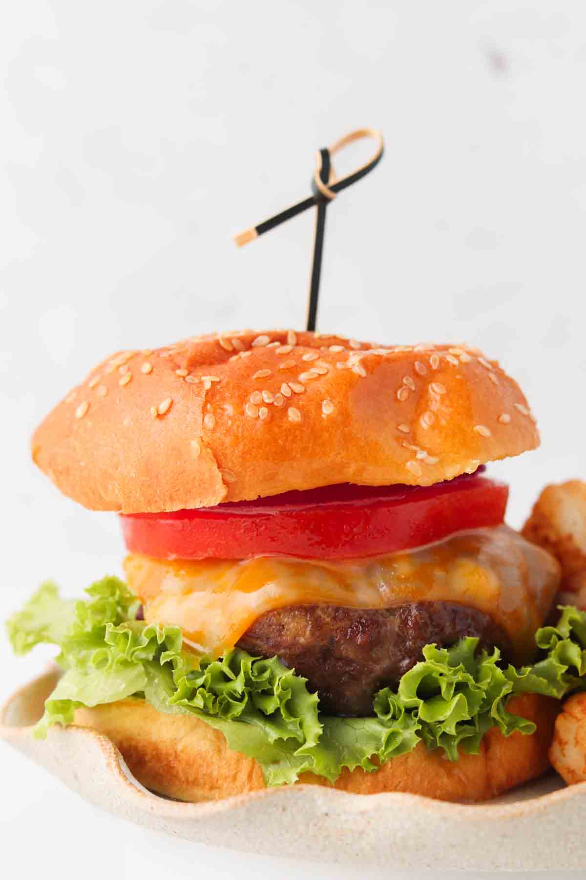 Gluten free beef burger with a bun on it with cheese, tomato and lettuce on a plate with tater tots.