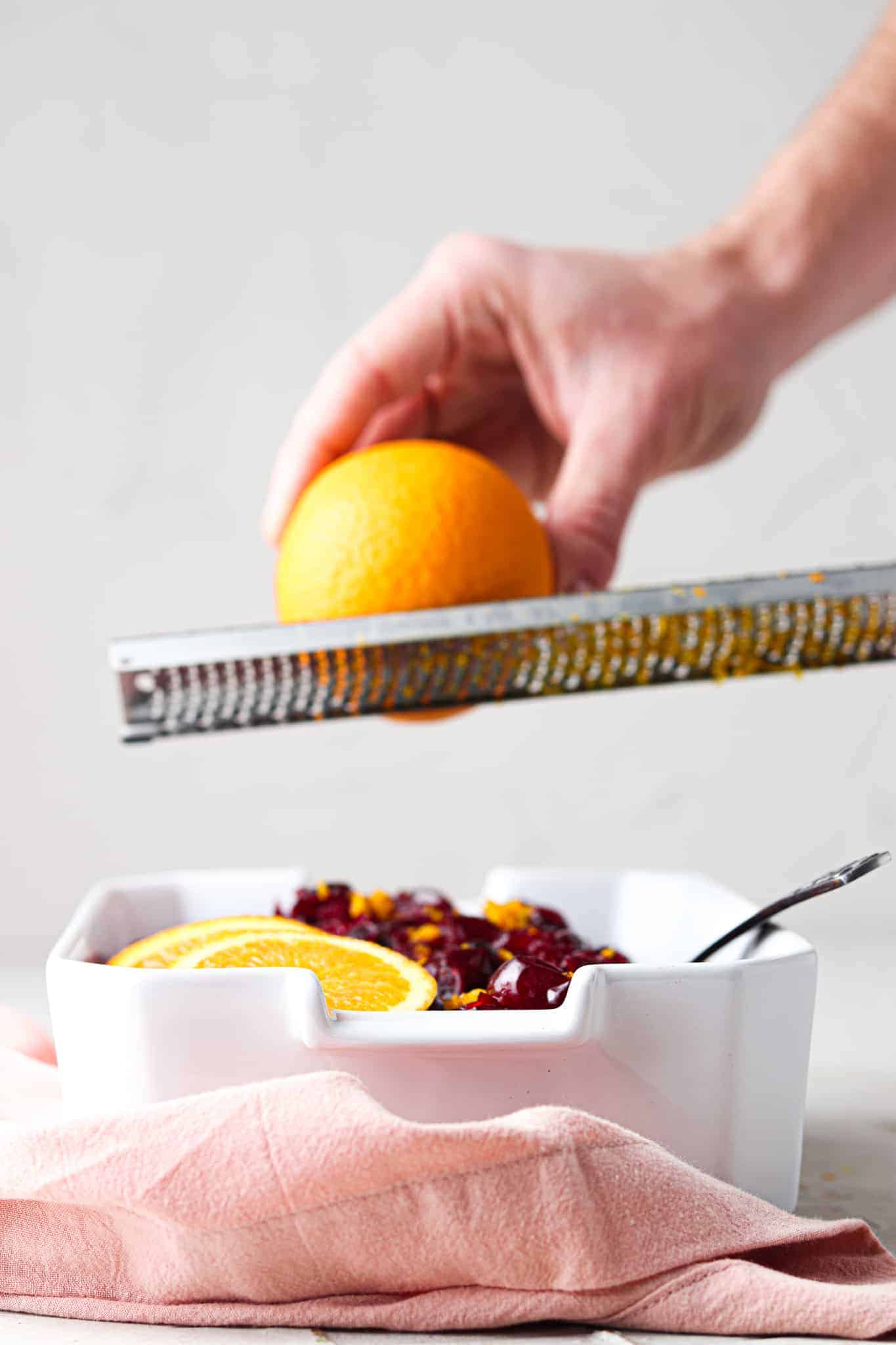 Hand holding an orange zesting it over a lemon zester over a white casserole dish filled with cooked cranberry sauce.