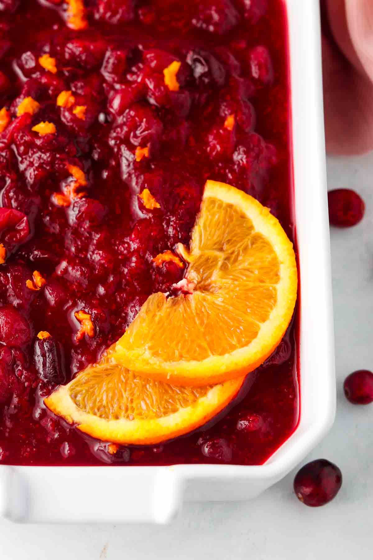 Cooked cranberry sauce garnished with orange zest and orange sliced in a white baking dish.