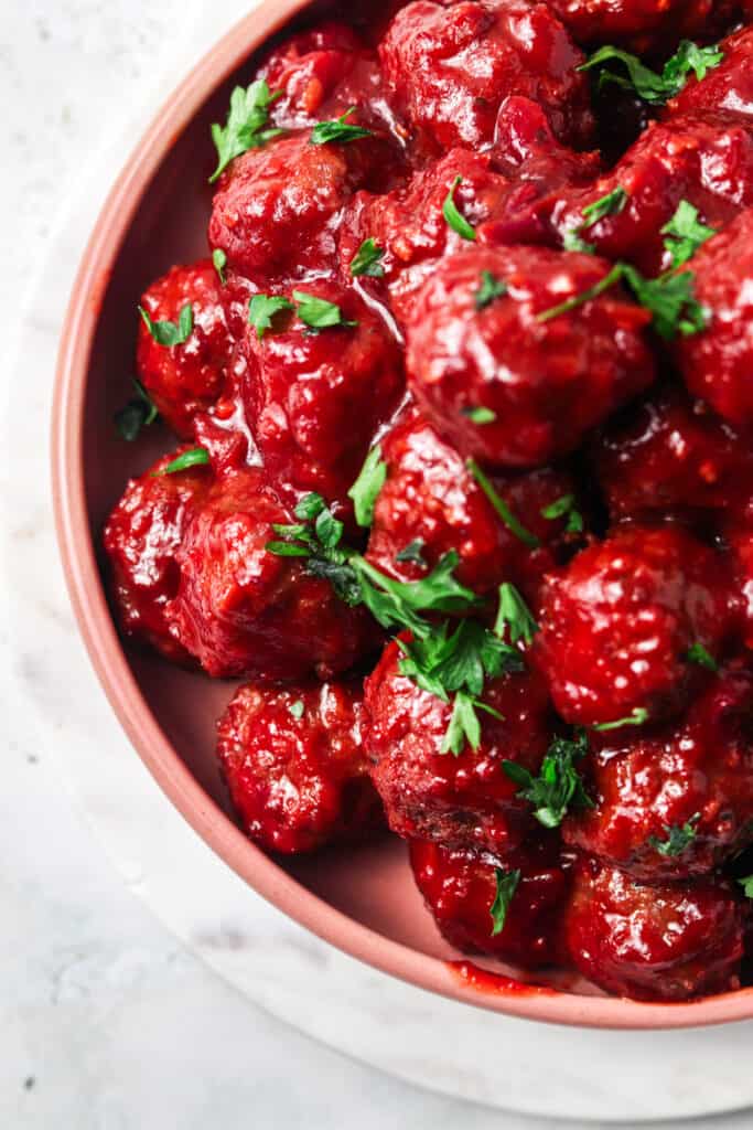 Paleo cranberry bbq meatballs in a salmon colored serving dish on a white marbled tabled and garnished with fresh green parsley.