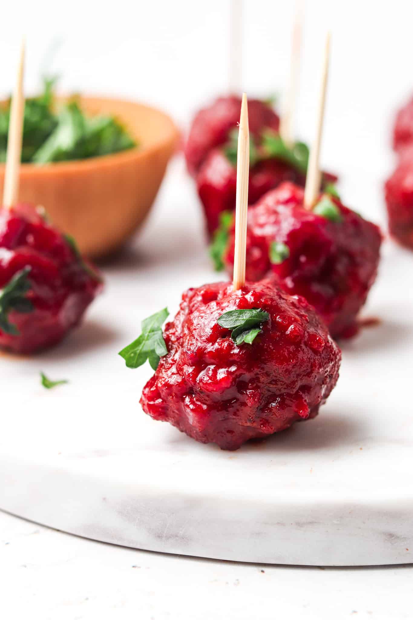 Paleo cranberry bbq meatballs on a white platter with a wooden toothpick in it and garnished with fresh green parsley.