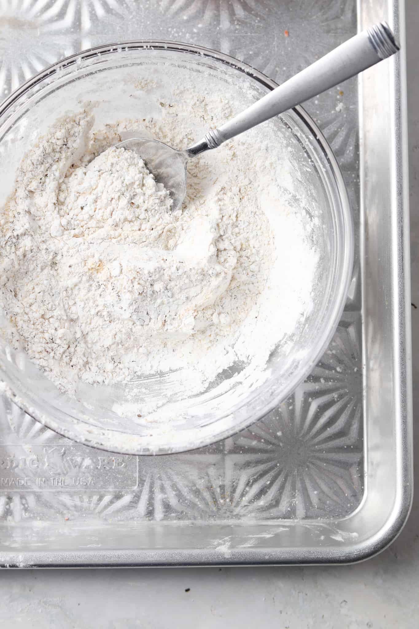 Arrowroot powder, garlic powder, onion powder, dried oregano, dried basil, and sea salt all mixed in a clear mixing bowl on a silver backing sheet with a silver spoon in the bowl.