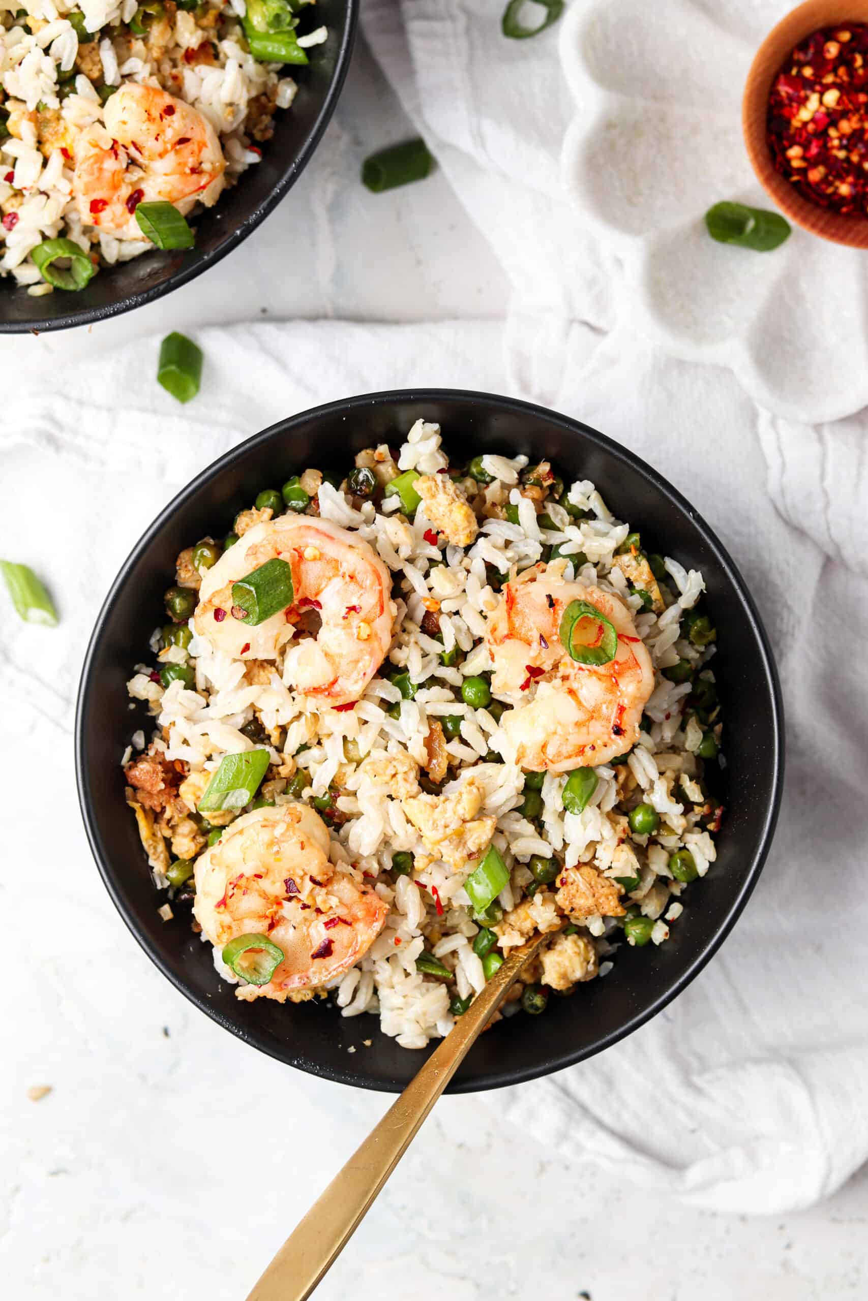 Shrimp fried cauliflower rice in a black bowl with a spoon and red pepper flakes.