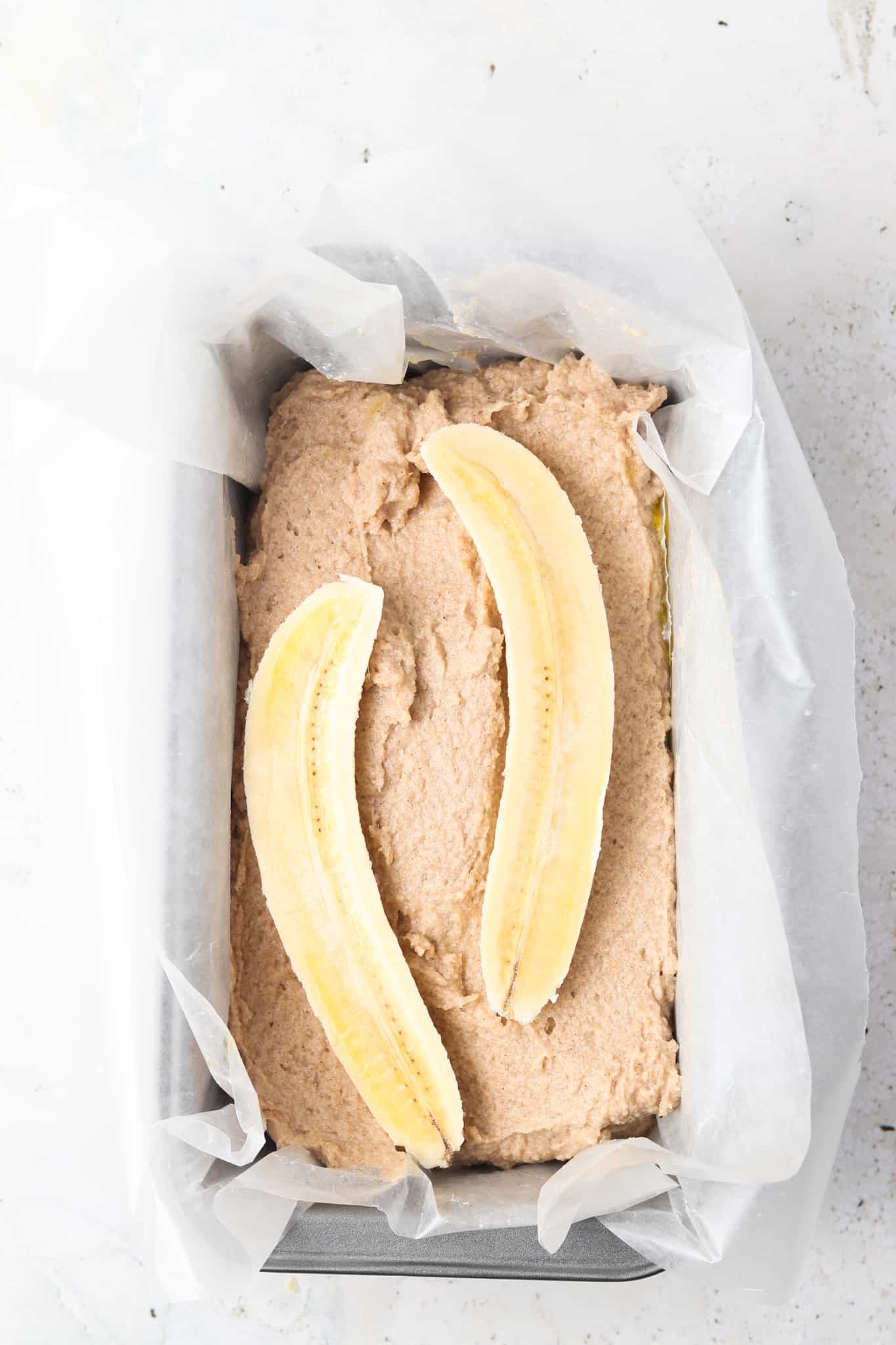 Banana bread mixture in a loaf pan lined with wax paper and topped with halved banana. 