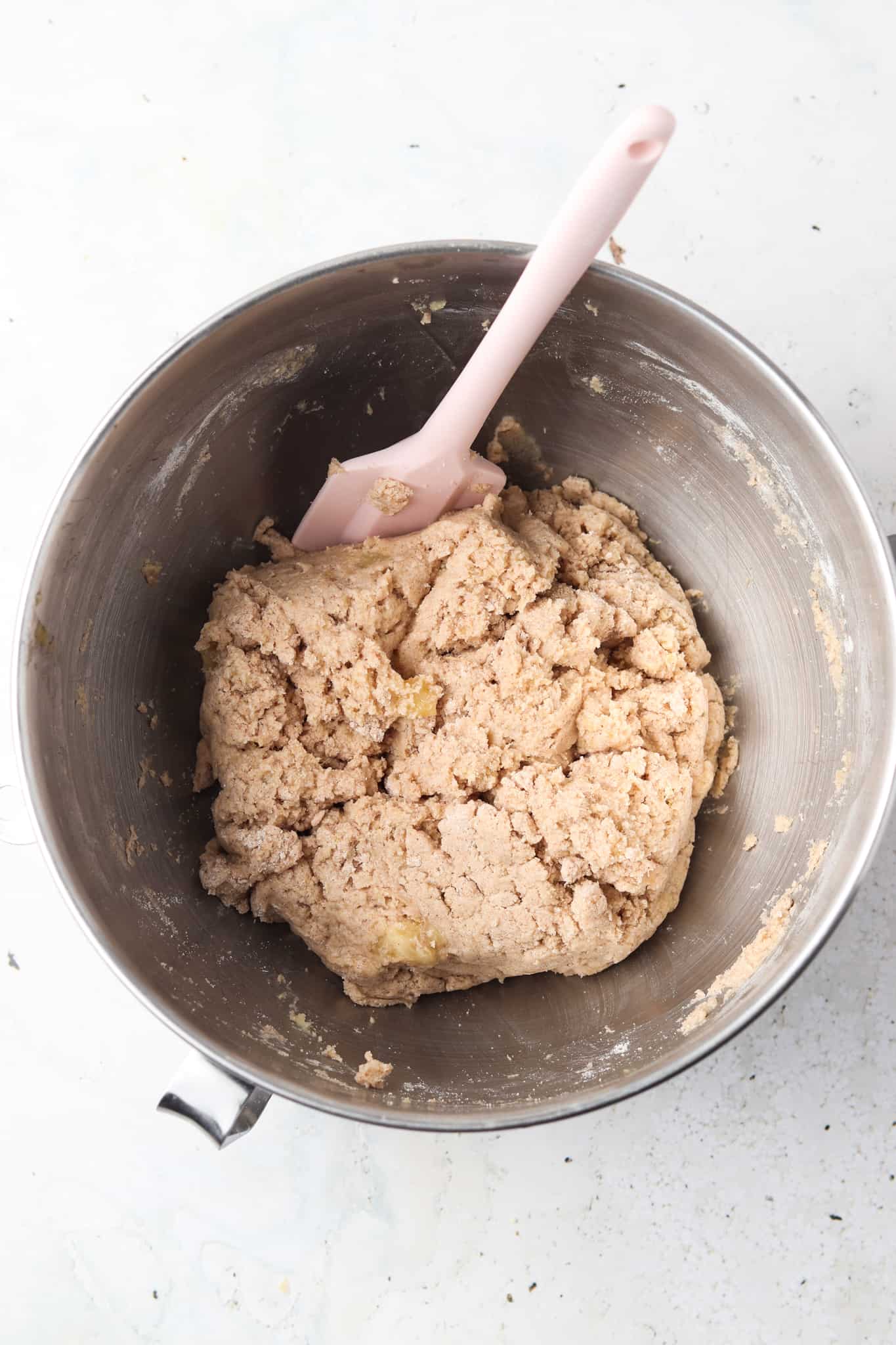 Bananas, cassava flour, arrowroot flour, coconut flour, applesauce, maple syrup, ground ginger, cinnamon, vanilla powder, baking soda, baking powder, and extra virgin olive oil in a silver mixing bowl with a pink spatula in it.