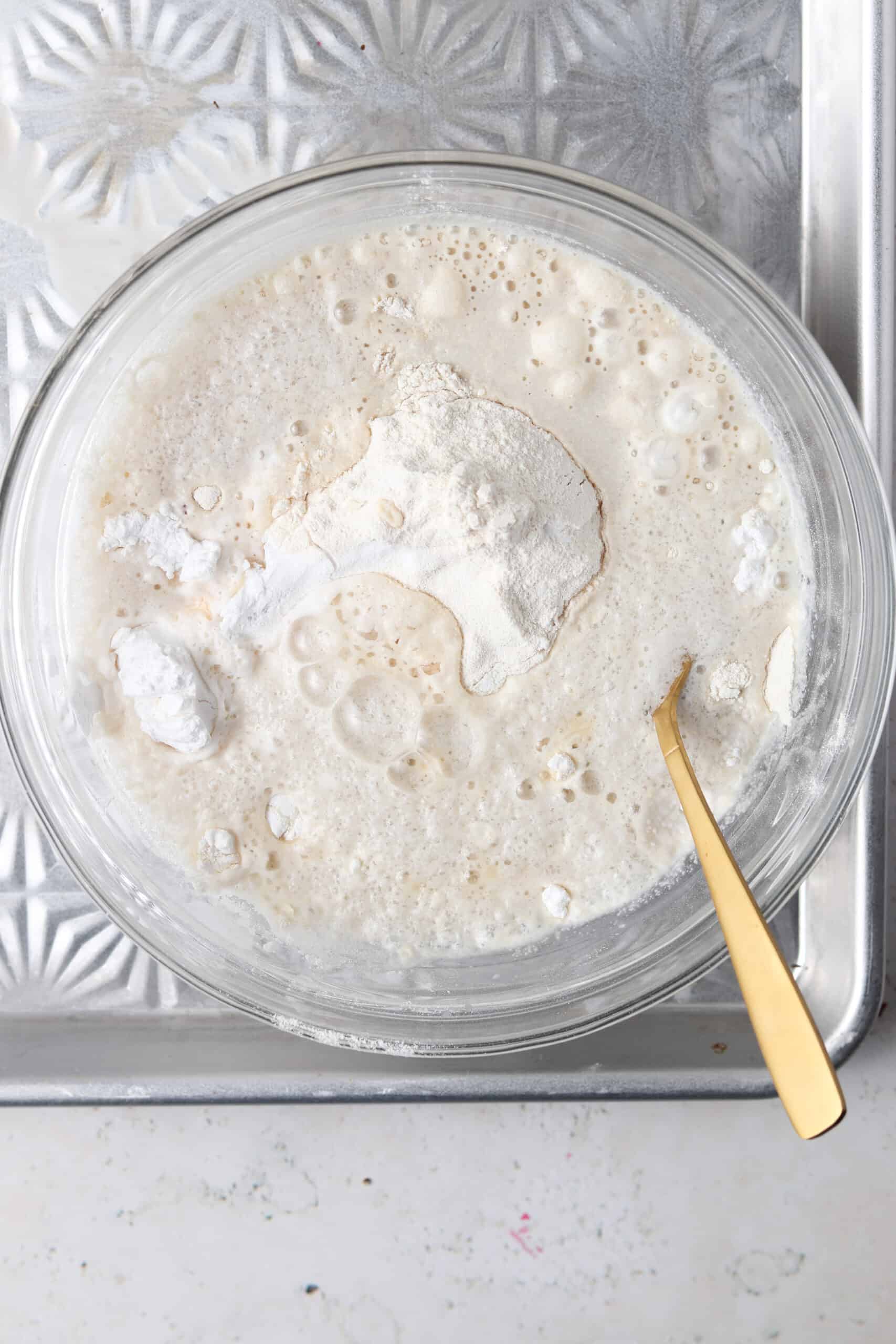 Cassava flour, tapioca flour, baking powder, banana, coconut milk, vanilla extract, cinnamon, pure maple syrup, and water in a glass mixing bowl on a silver platter with a gold fork in it.