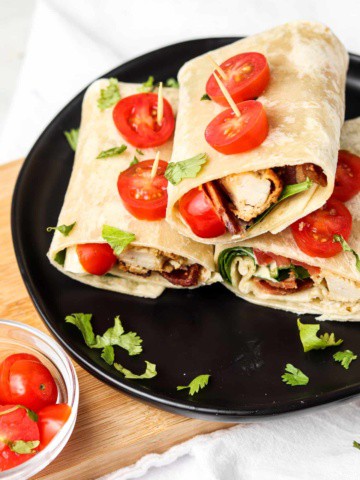 crispy bacon chicken ranch wraps on a black plate with slices of tomato held on with toothpicks