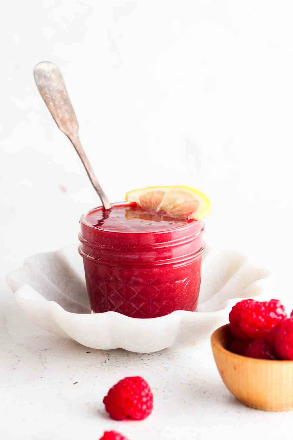 Raspberry jam in a small mason jar with a lemon on top and raspberries laid next to it.