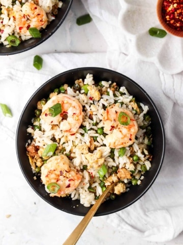 Shrimp fried cauliflower rice in a bowl with green onions and red pepper flakes on top.