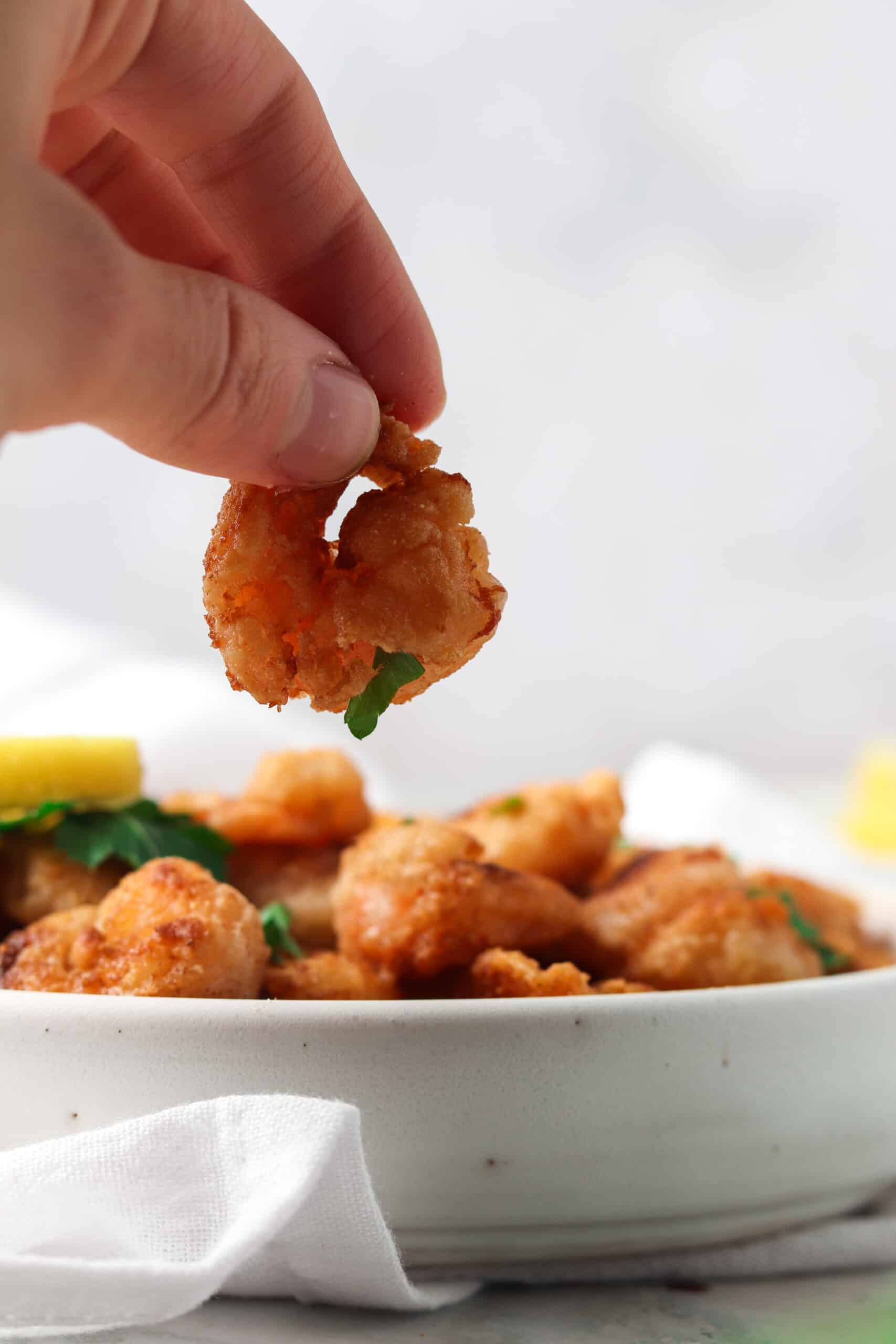 Person holding a piece of gluten free fried shrimp over a white plate.