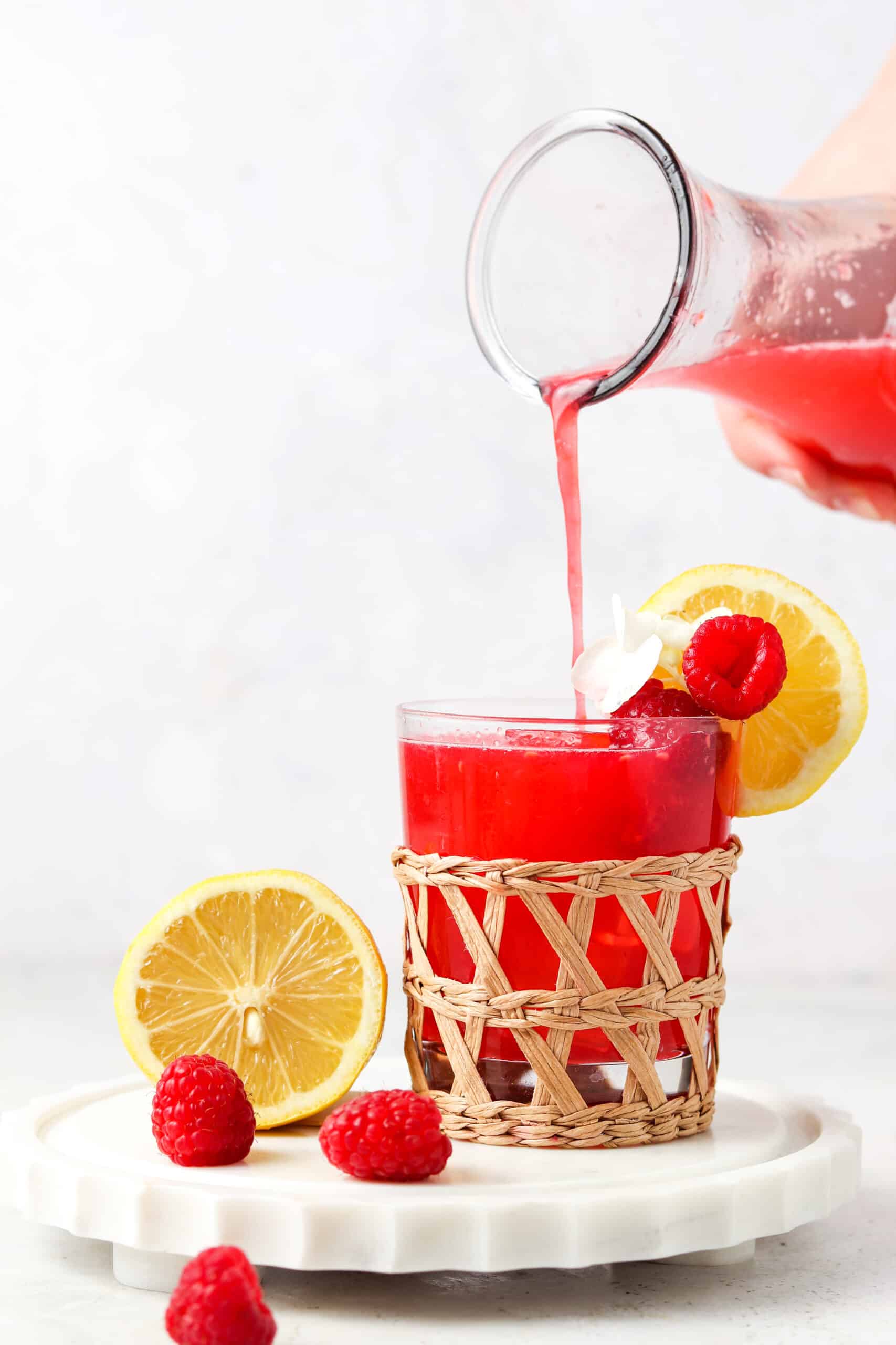 Red colored drink in a large mason jar being poured into a small whicker glass with raspberries on the side. 