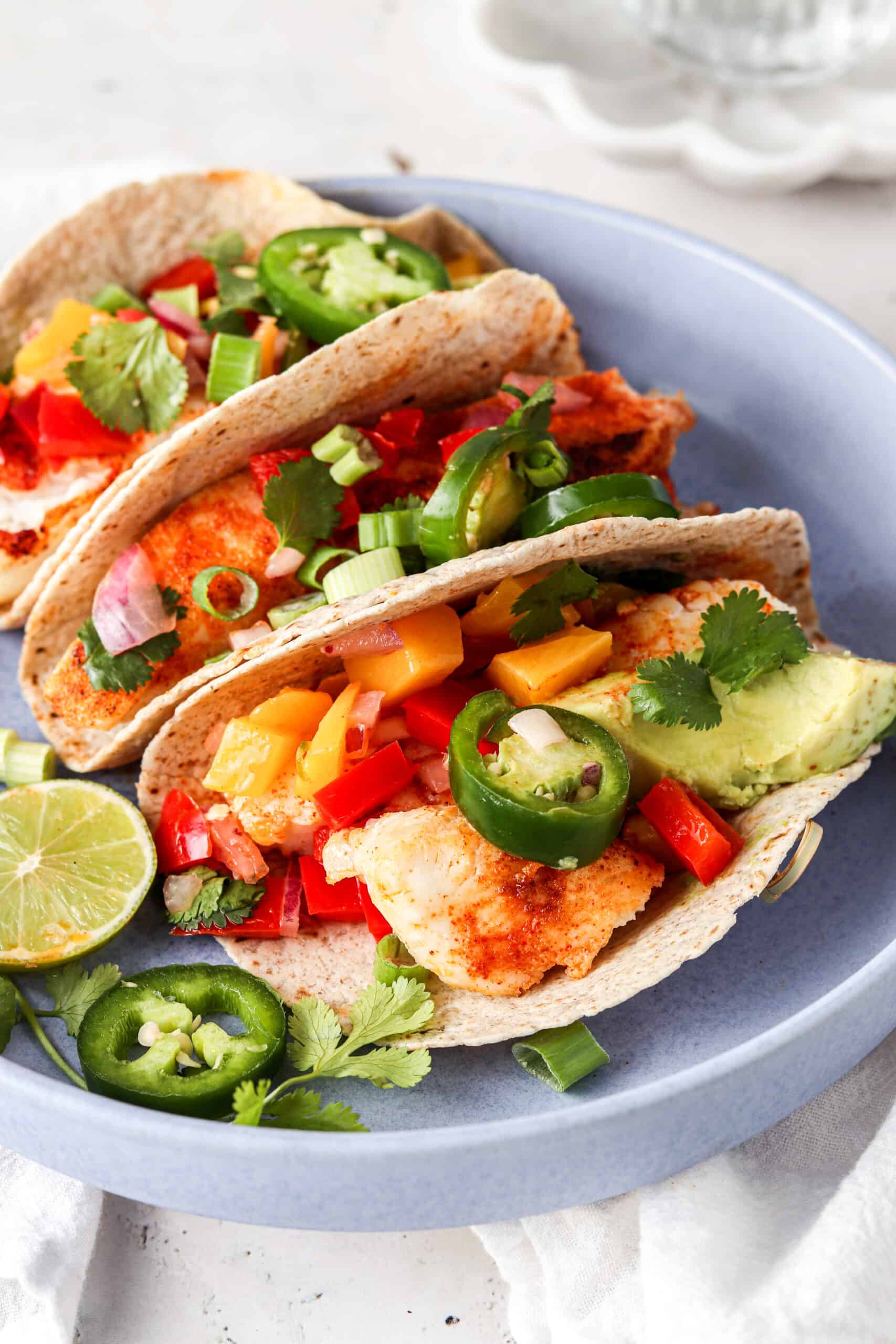 Halibut fish tacos lined up in a blue bowl and garnished with fresh green herbs and jalapeños.