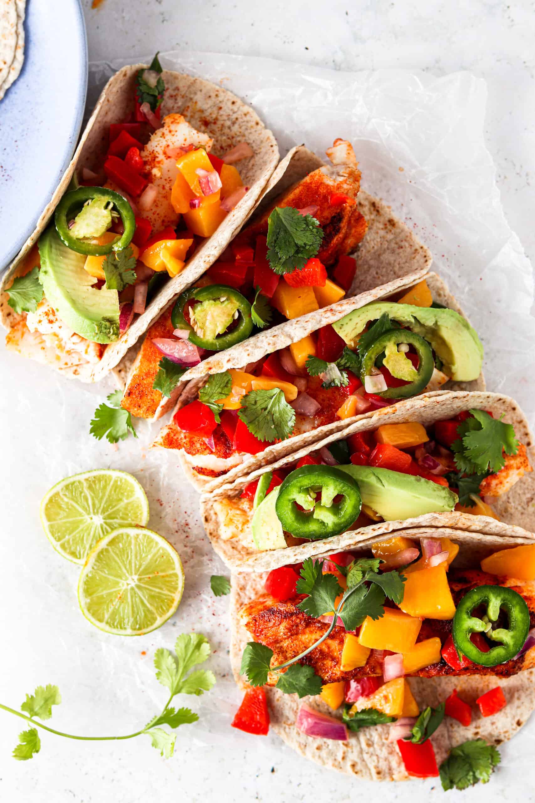 Halibut fish tacos in a line on a platter with fresh cilantro, avocado, and jalapeño slices. 