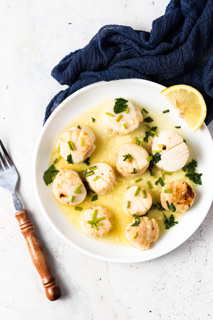 whole 30 scallops with ghee, garlic, and chives on a white plate with lemon wedge and a navy blue fabric next to it and a fork