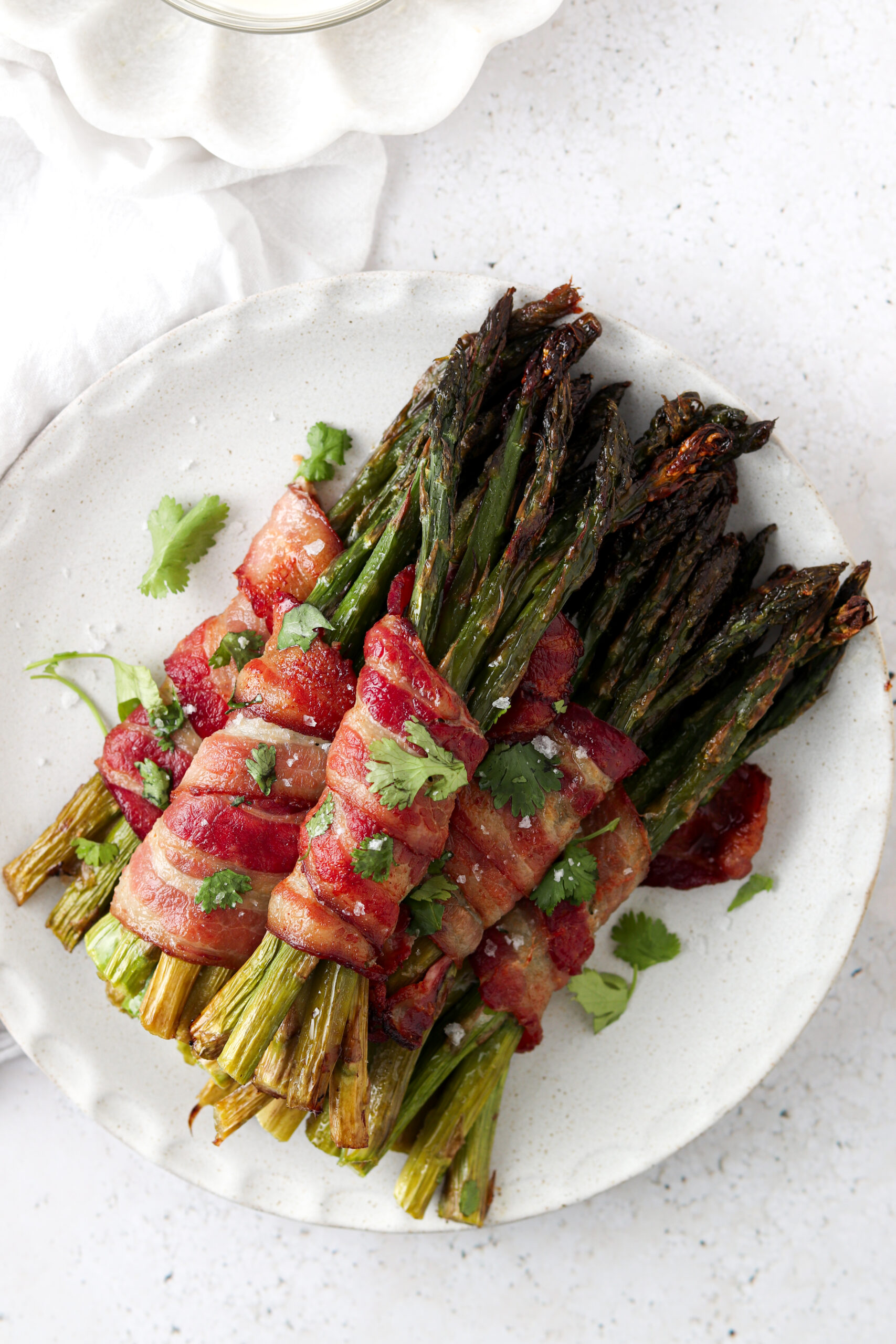 Bacon wrapped asparagus stacked on a white plate and garnished with fresh green herbs.