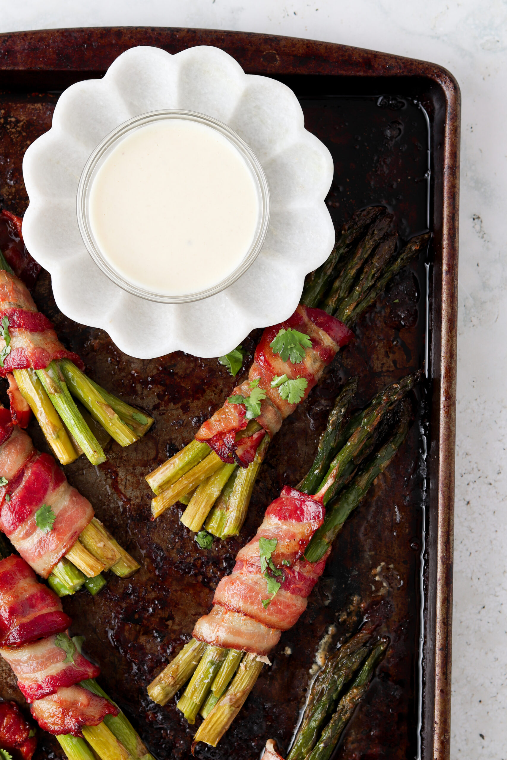 Cooked bacon wrapped asparagus bunches on a baking sheet with garlic aioli in a small bowl. 