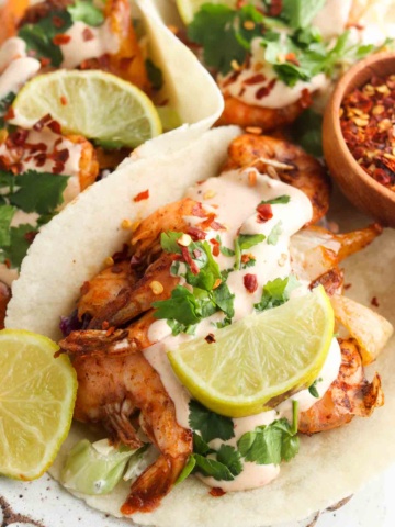 Close up photo of shrimp tacos on a plate with limes, coleslaw and spicy mayo.