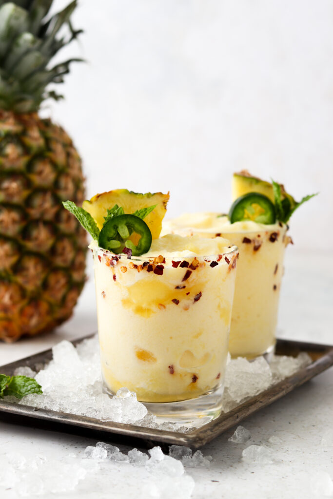 Two pineapple margarita mocktails on a platter with ice and jalapenos on top.