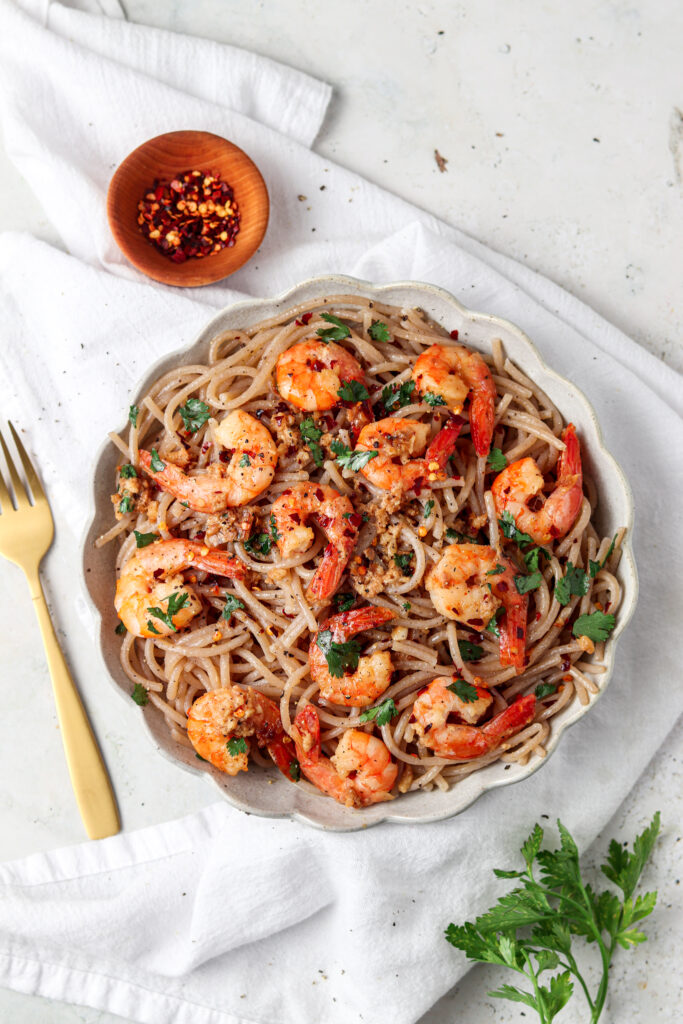 gluten free noodles with shrimp scampi on a white plate with a gold fork a side of red pepper flakes on top of white fabric