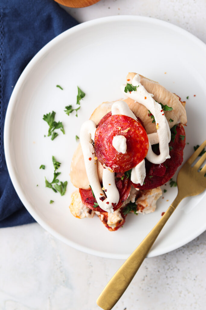 pizza chicken on a white plate with parsley sprinkled on top and a slice of pepperoni on top with a gold fork