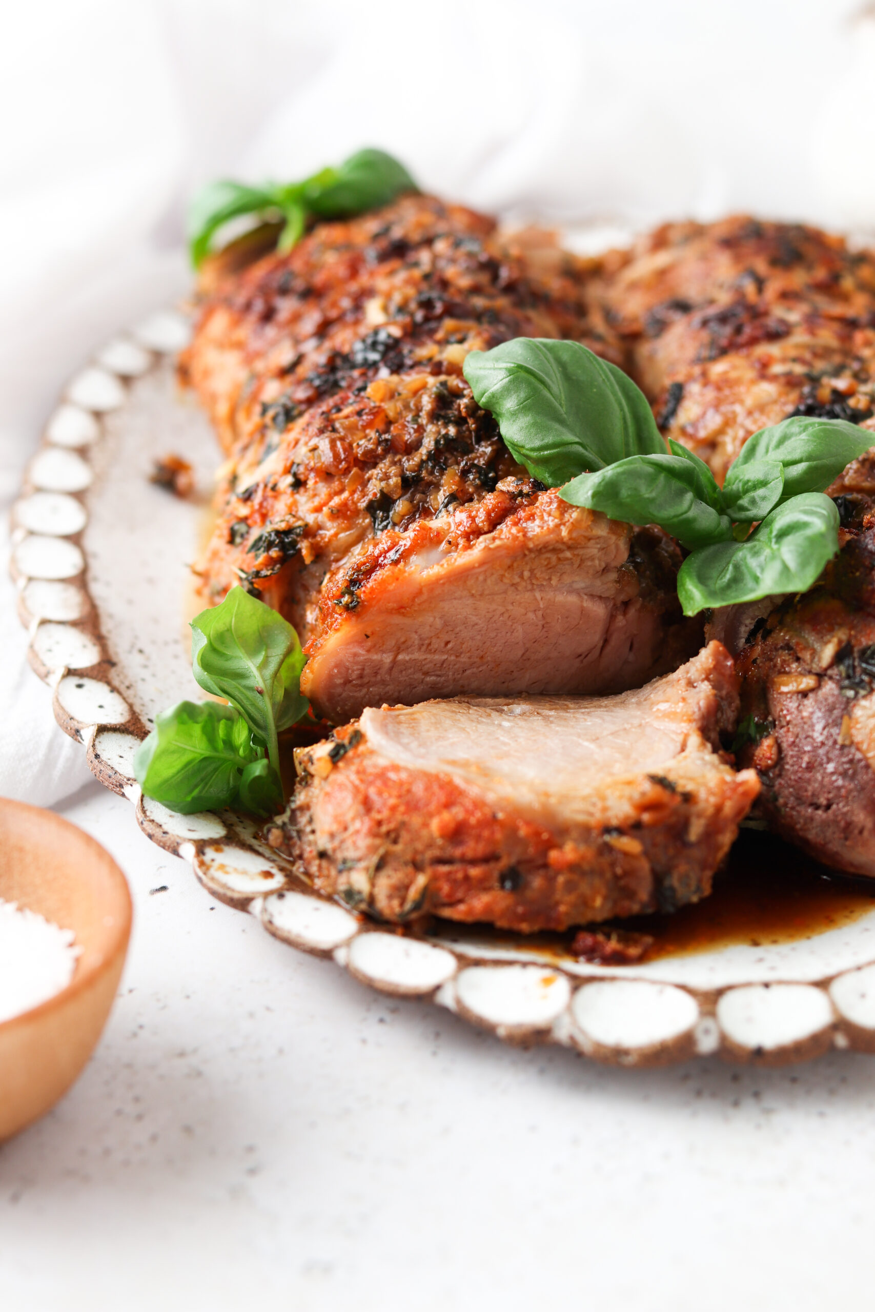 Whole30 pork tenderloin sliced on a platter with fresh green basil as a garnish on a white surface. 