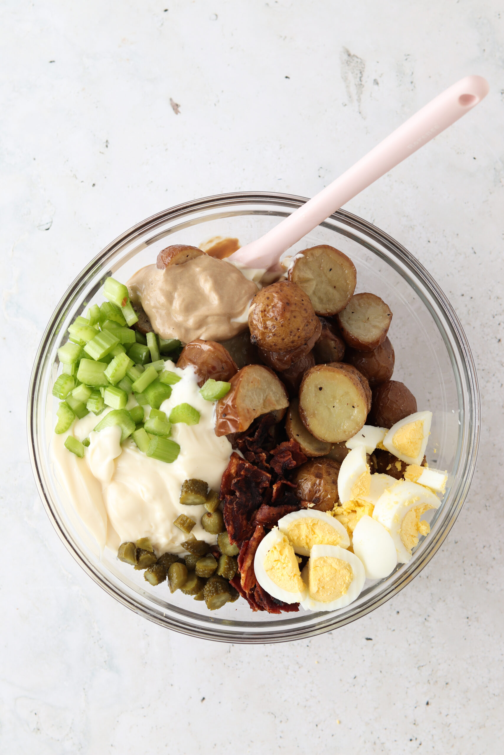 Mini potatoes, eggs, bacon, celery, pickles, and mayo in a glass container with a spoon. 