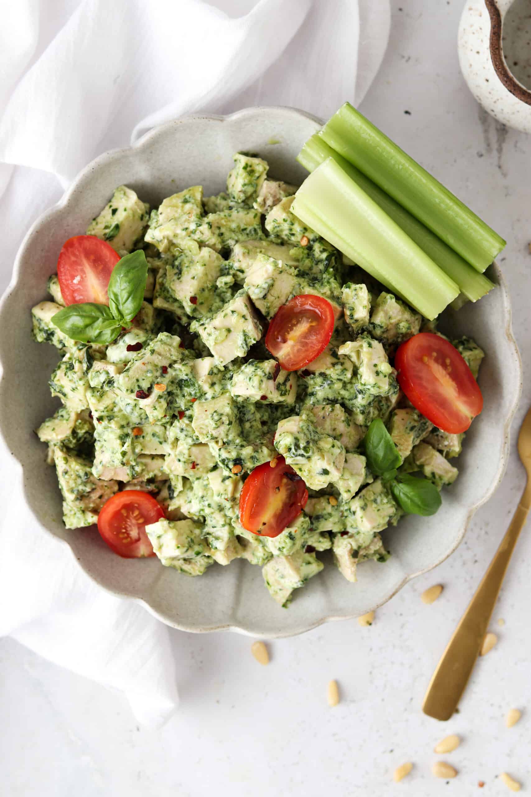 Pesto chicken salad in a bowl with celery sticks and halved grape tomatoes. 