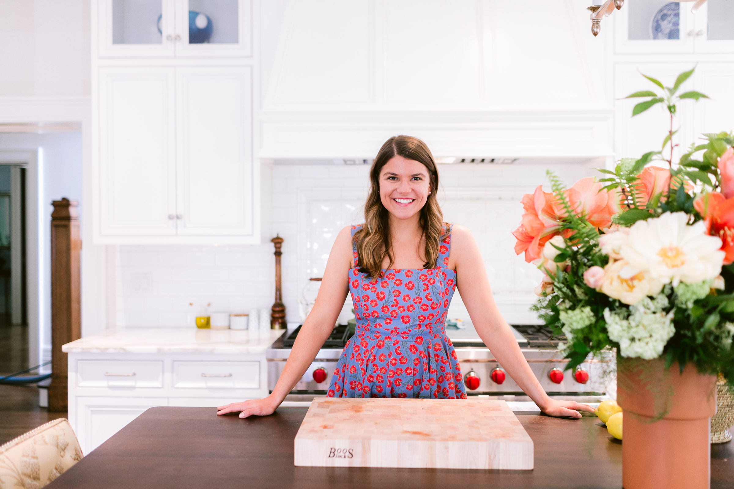 Picture of a girl in a red and blue dress in a kitchen with a wood cutting board. 