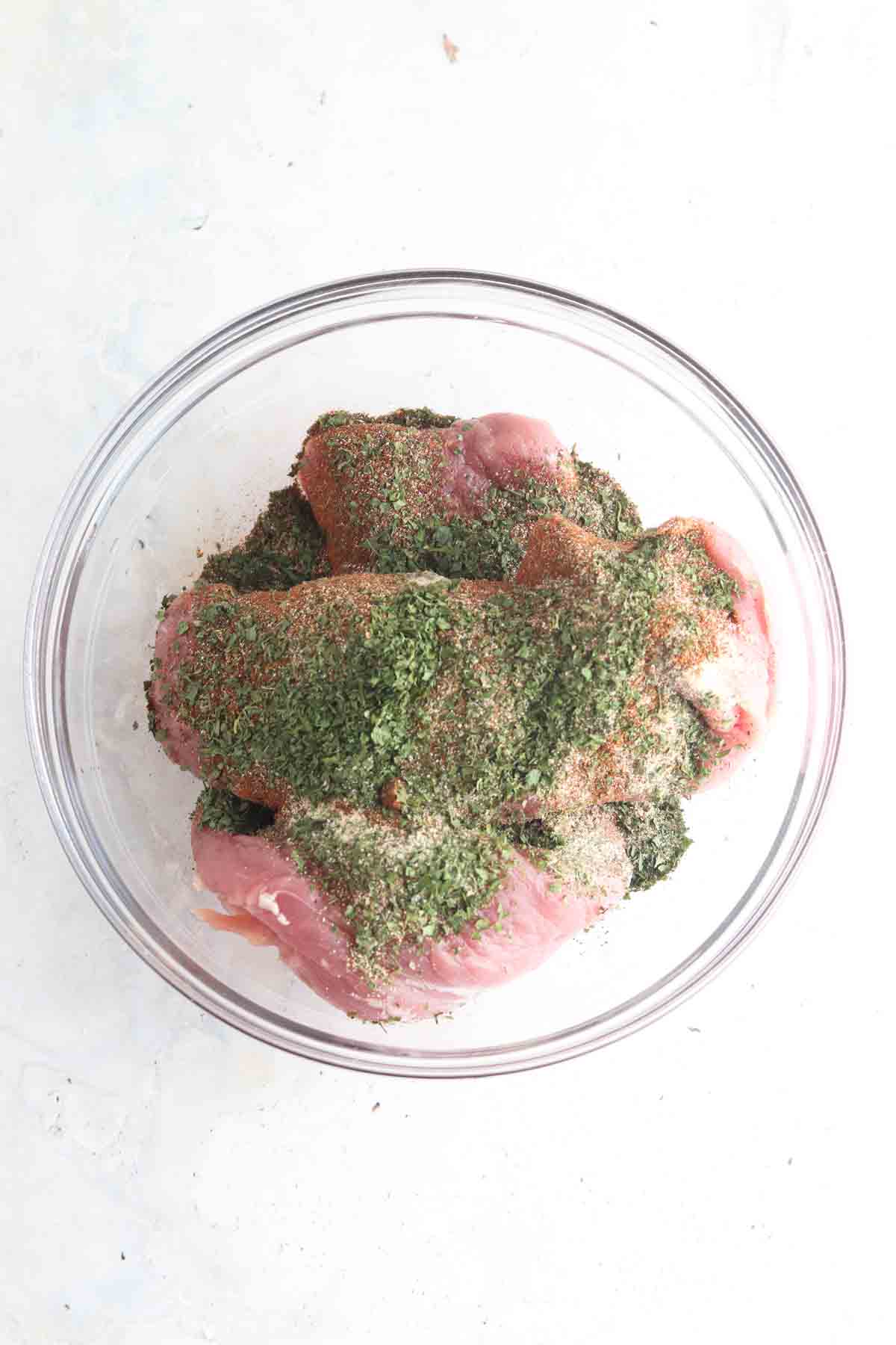 Raw pork tenderloin in a bowl with seasoned herbs on top.