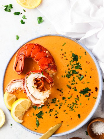 dairy free lobster bisque in a bowl with a gold spoon and garnished with lemon, fresh green herbs, and red pepper flakes.