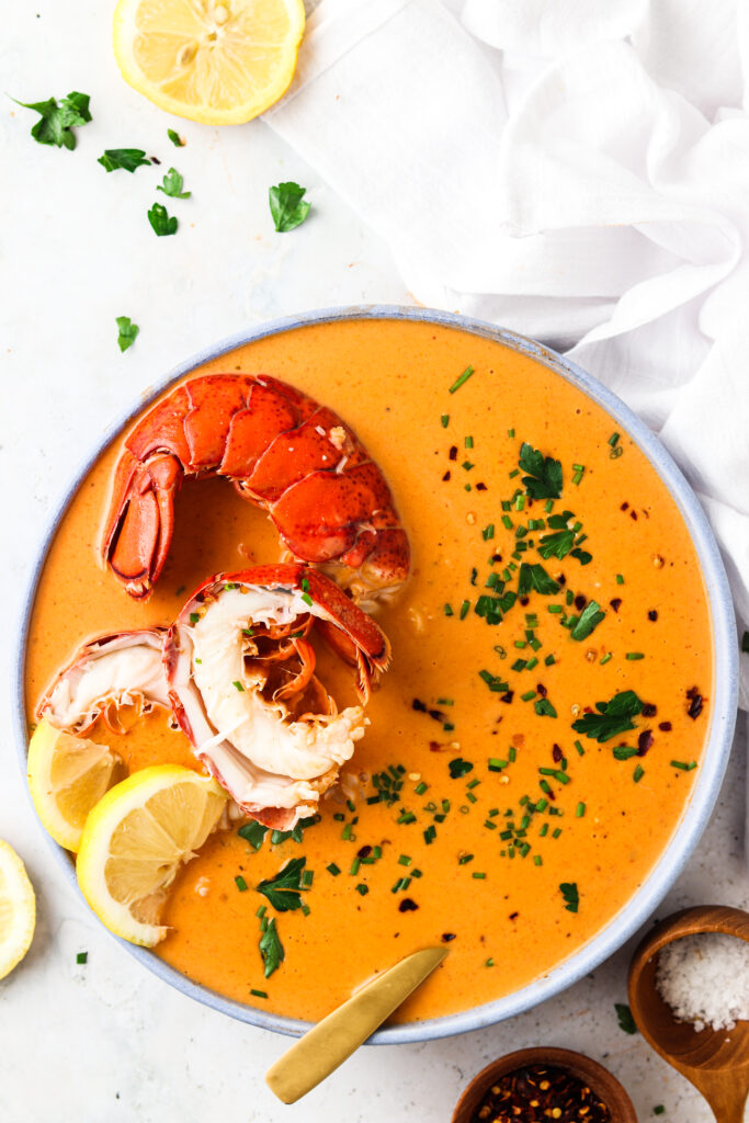 dairy free lobster bisque in a bowl with a gold spoon garnished with a lobster tail, lemon, fresh green herbs, and red pepper flakes