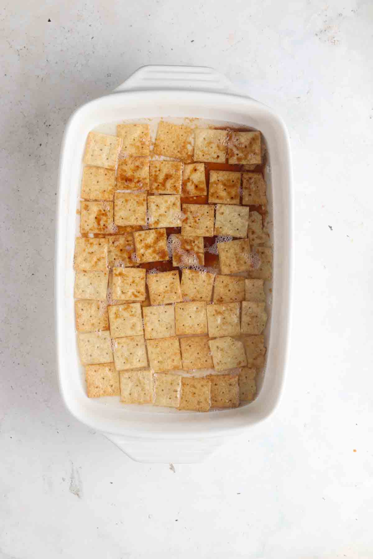 Almond flour crackers in a white casserole dish with melted coconut oil, maple syrup, and coconut sugar mixture poured overtop. 