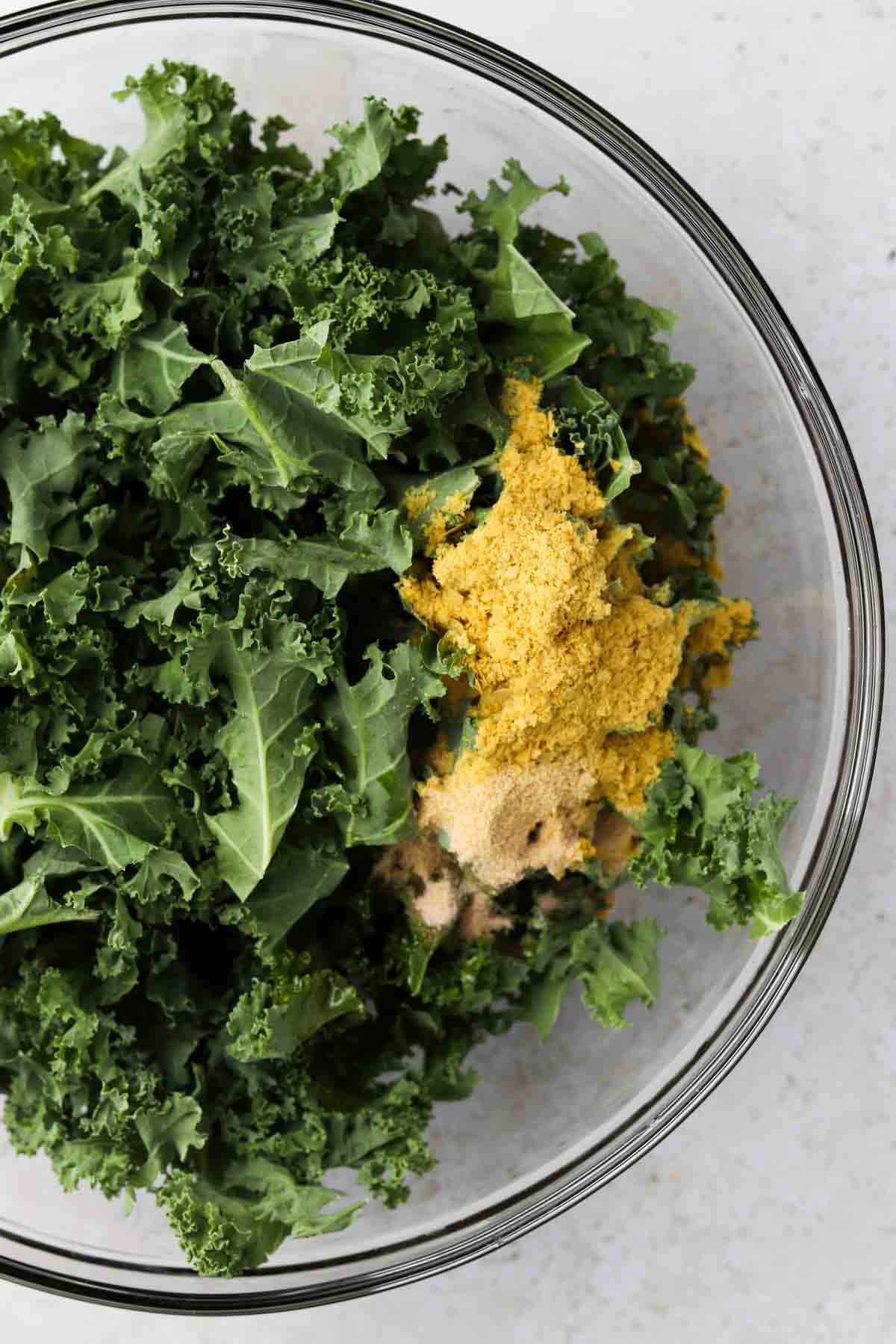 Kale, nutritional yeast, sea salt, garlic powder, and avocado oil in a glass mixing bowl.