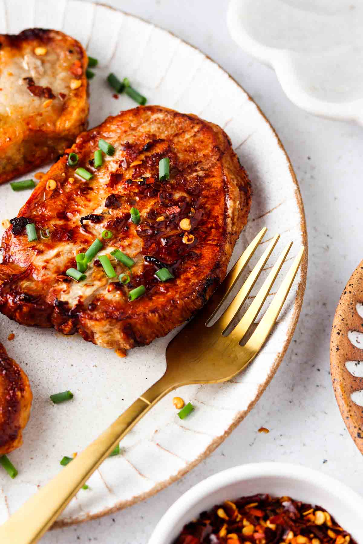 Cooked pork chops on a white plate with a gold fork and garnished with chives and red pepper flakes.