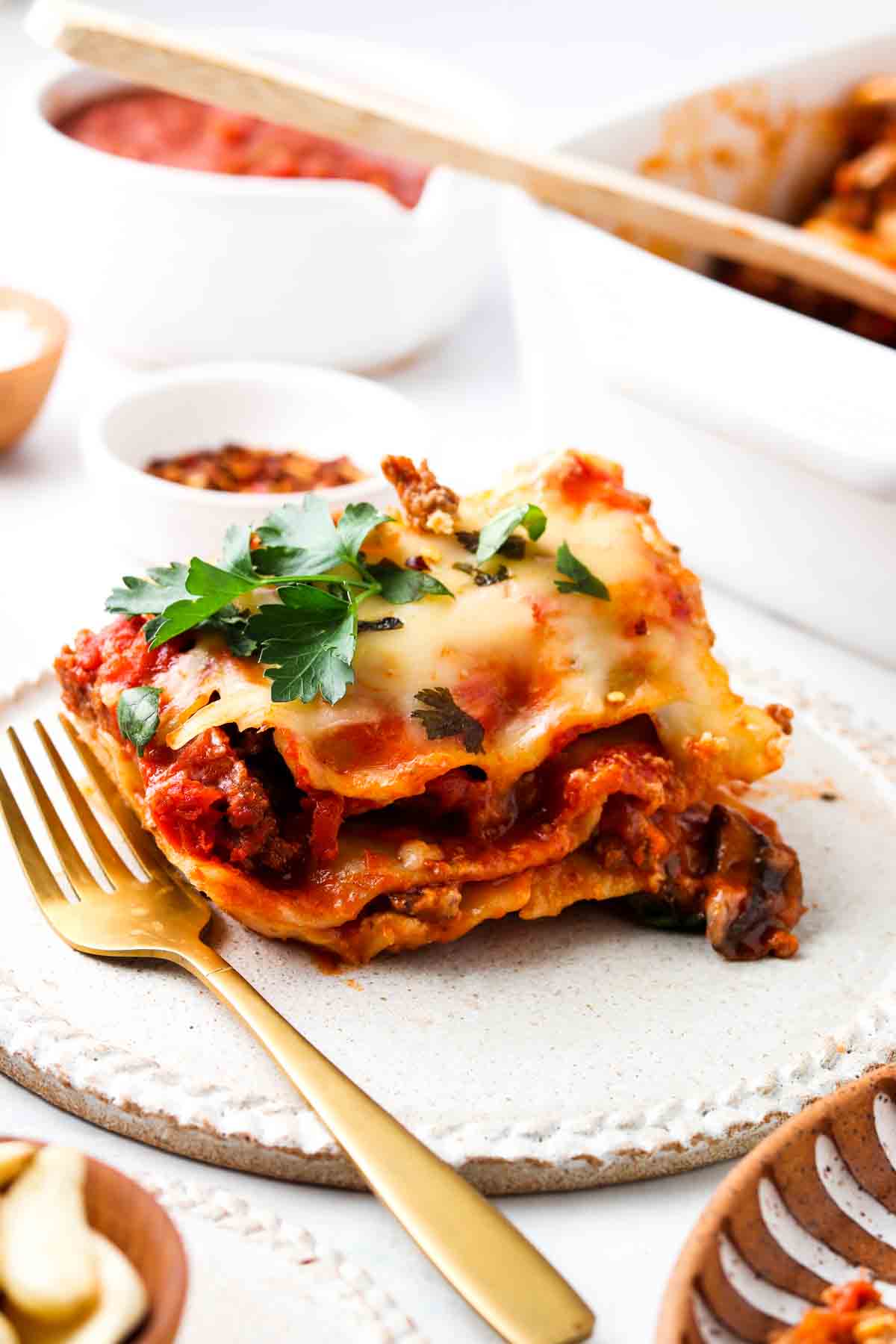 Piece of lasagna with gluten free noodles and cheese on a plate with a fork and fresh herbs on top.